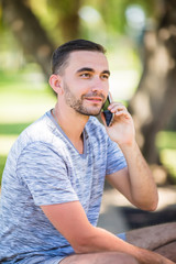 Handsome young man talking on phone while sitting on bench in park