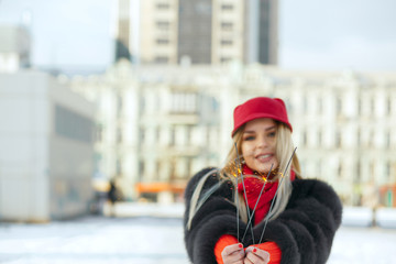 Cool blonde girl wearing fashionable red cap and scarf celebrating Christmas with sparklers at the street. Space for text