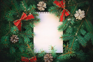 Fototapeta na wymiar Top view of christmas new year wishes notebook paper card note with frame of fluffy needless spruce tree, bows, pine cones. Flat lay.
