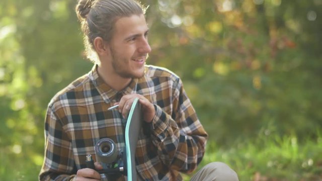 Portrait of a young smiling guy in a plaid shirt customizing a skateboard. Young man with a skateboard outdoors.