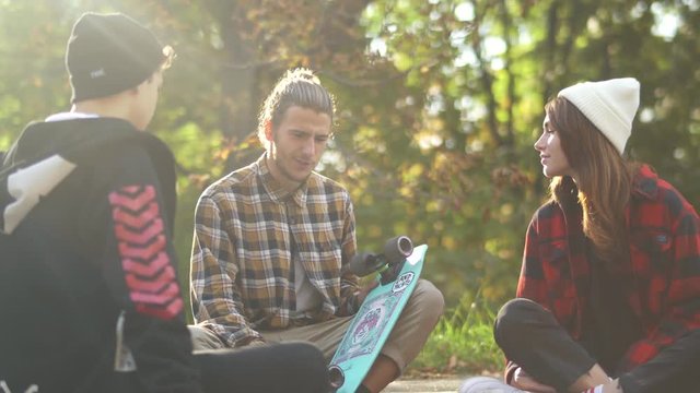 Company of hipsters with skateboard sitting in the park. Friends chatting outdoors. A young guy in a plaid shirt with a skateboard sitting among friends. The girl in a plaid shirt and in a white hat.