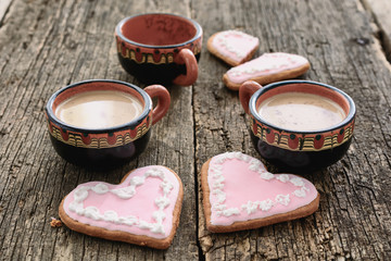 Two full cups of coffee next to two heart-shaped gingerbreads are close to each other and one empty coffee cup next to a broken heart-shaped gingerbread at a distance on old wooden boards