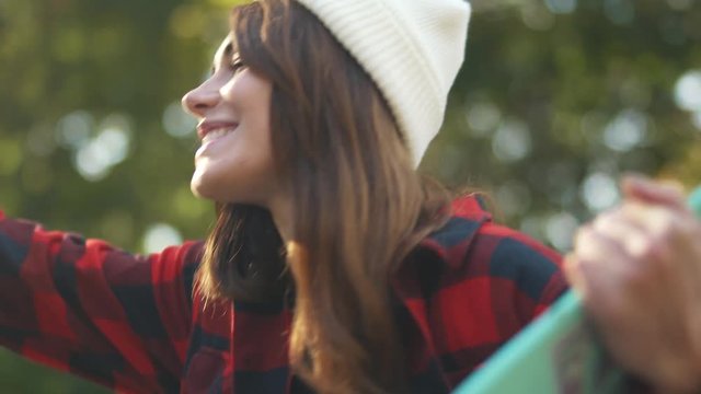 The guy passes the skateboard to the girl in a plaid shirt and white hat. Girl takes a skateboard and hugs her friend while sitting outdoors.