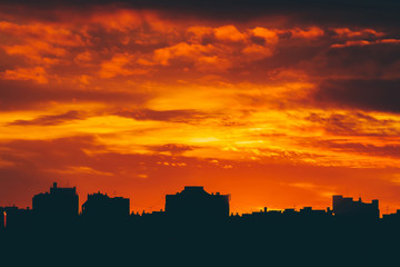 Naklejka premium Cityscape with vivid fiery dawn. Amazing warm dramatic cloudy sky above dark silhouettes of city buildings. Orange sunlight. Atmospheric background of sunrise in overcast weather. Copy space.