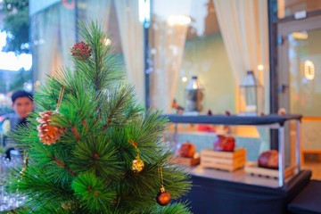 Small green Christmas tree with pine cones, sparkling gold and red ball ornaments and decoration. Happy New Year festive celebration. Season Greeting. Window display city background. Natural light.