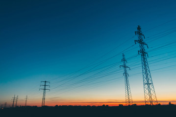 Power lines in field on sunrise background. Silhouettes of poles with wires at dawn. Cables of high voltage on warm orange blue sky. Power industry at sunset. Many cables in picturesque vivid sky.