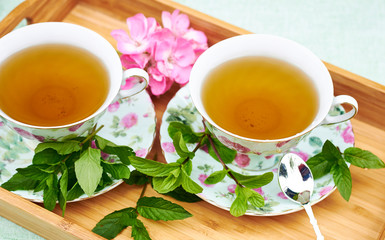 Morning herbal mint tea served on a tray: two cups with drink, fresh bunch of herb and a pink geranium flower on the background, view from above, closeup, copy space, crop. Romantic breakfast concept