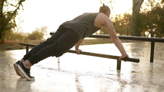 Sport, fitness, young muscular man working out during a workout on the street.Caucasian athlete making push ups on paralel bar. Morning sunshine on the background. Rare view