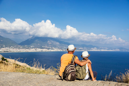 Young father and son with backpacks are sitting on the beach against the backdrop of the sea. Family travel concept