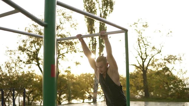 Handsome man having calisthenics training on horizontal bar outdoors. Does gymnastic stunts on the crossbar in the park. Lens flares on the background. Slow motion