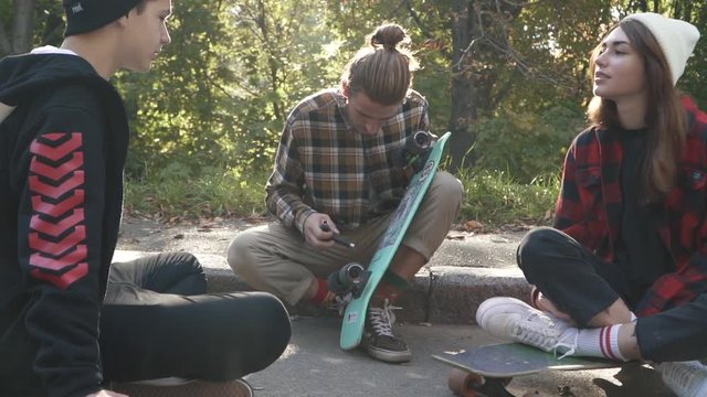 Teenagers are sitting with skateboards next to each other outdoors. Hipster friends chatting while sitting on a skateboard in the park. Young guy in a plaid shirt adjusts his skateboard.