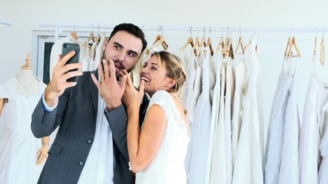 Romantic wedding couple in expensive dress shop. White man and woman in bride dress, video call to their friends on mobile phone. Romantic young couple concept.
