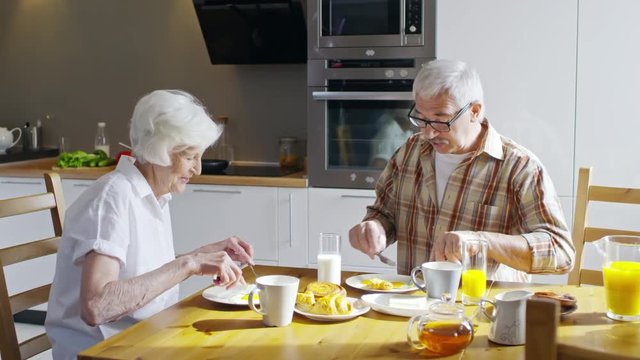 Medium shot of old couple sitting at breakfast table eating scrambled eggs while smiling and talking with each other