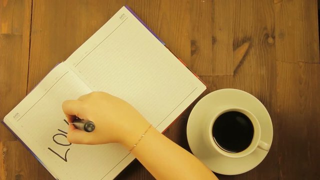 The girl writes in the diary the love word with a black marker and drinks coffee