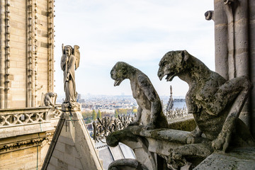 Two stone statues of chimeras overlooking the rooftop of Notre-Dame de Paris cathedral from the towers gallery with the statue of an angel with trumpet and the city vanishing in the mist.