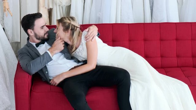 Romantic wedding couple in expensive dress shop. White man and woman in bride dress, laying on red sofa being intimate. Romantic young couple concept.