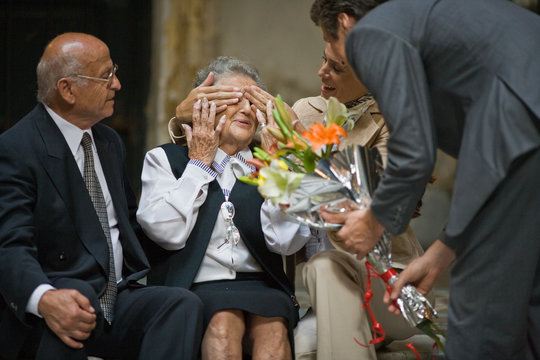 Mid-adult woman covering her senior mother's eyes as her brother presents a bouquet of flowers while sitting near her senior father.