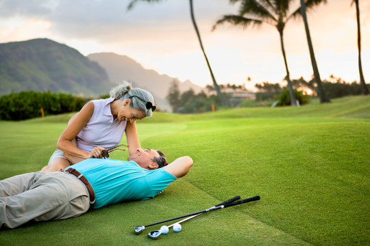 Smiling female golfer sits on the green and leans over a smiling male golfer lying on the ground next to her with his arm around her.