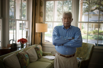 Portrait of a mature adult man standing with folded arms in a living room.