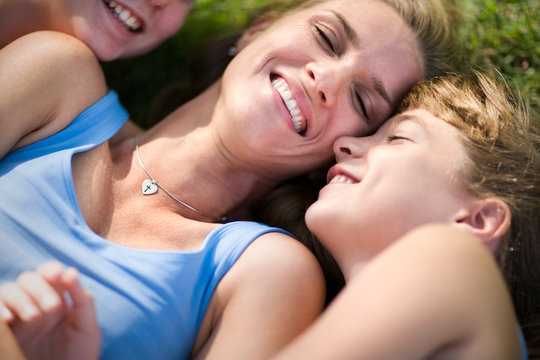 Mid-adult woman lying on the grass being hugged by her daughters.