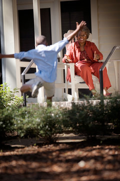 Mature adult woman sitting on a porch watching her young grandson jump over some shrubs.