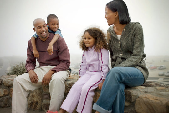 Happy mid-adult couple and their two young children sitting on a stone wall outdoors.