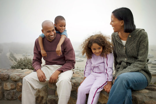 Happy mid-adult couple and their two young children sitting on a stone wall outdoors.