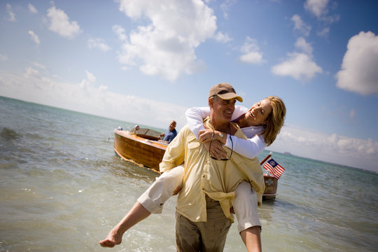 Smiling mature man piggybacking his wife from a boat to the shore on a beach.