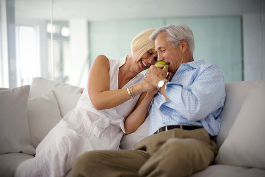 Mature man biting into an apple held by his wife as they sit together on couch in their living room.
