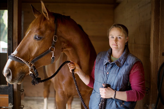 Portrait of a young woman standing with her brown horse inside a stable.