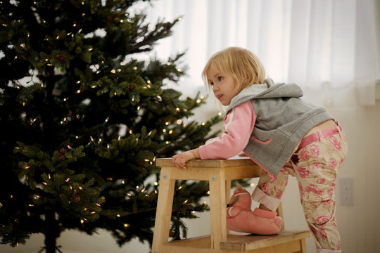 Young girl climbing a stool next to a Christmas tree.
