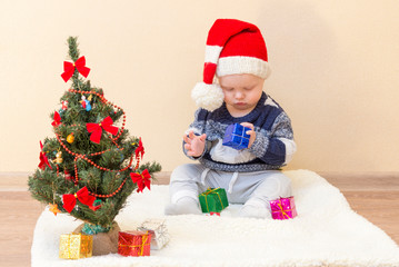 Funny baby in Santa Claus hat sitting near the Christmas tree with gifts