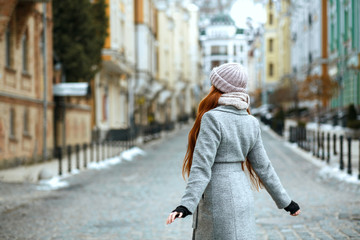 Pretty red head woman wearing winter cap and coat posing at the city. View from the back. Empty space