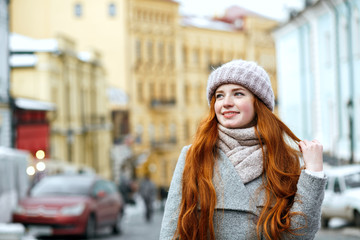 Street portrait of joyful redhead girl with long hair wearing warm winter apparel posing at the street. Empty space