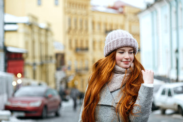 Naklejka na ściany i meble Street portrait of lovely redhead girl with long hair wearing warm winter apparel posing at the street. Empty space