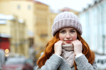 Closeup portrait of pretty red haired girl wearing knitted warm cap and scarf posing at the street. Empty space