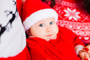 Close-up of a baby with blue eyes in Santa's costume between parents who looks at the camera lens