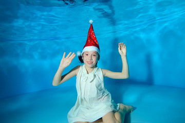 Obraz premium A little girl is sitting underwater at the bottom of the pool in a red Santa hat and a white dress on a blue background. She raised her hands up, looks at the camera and smiles. Portrait