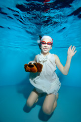 The child is a little girl, with a toy in her hand sitting underwater at the bottom of the pool in a red Santa hat, in a white dress and swimming goggles, and smiling on a blue background. Portrait