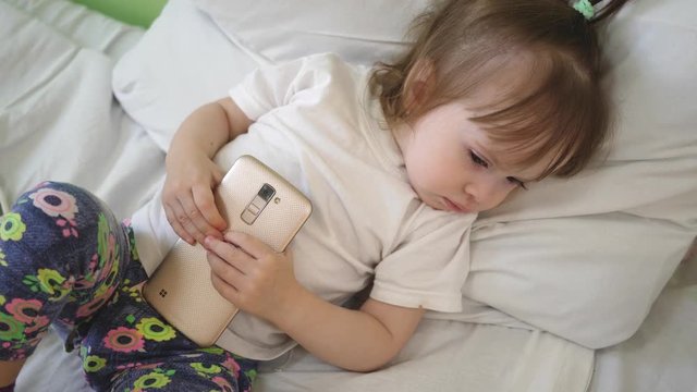 child is sleeping on pillow and holding tablet. Cute baby sleeping in bed with smartphone.