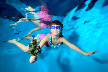 Cute little girl swims and poses under the water with a Christmas tree in her hand, in a pink...