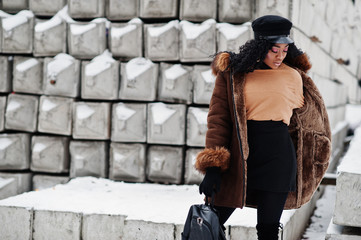 African american woman in sheepskin coat and cap posed at winter day against snowy stone background.