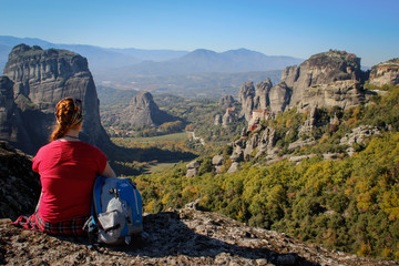 Naklejka premium A traveler girl in a red T-shirt and a blue backpack sits high against the rocks of the Meteora temple complex in Greece on a bright summer sunny day.