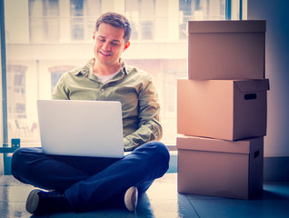 Handsome man with moving boxes and laptop sitting on stairs in house. European ethnicity