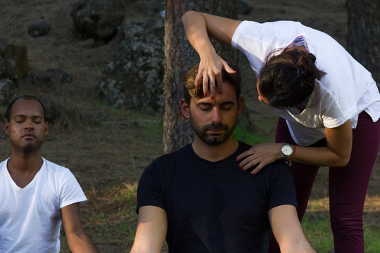 Mindfulness Female Teacher Applying Technique On Young Hipster Forehead In Forest Park. Yoga Meditation Class With Multi Ethnic Group Of Students In The Woods. Relaxation Exercise Concept
