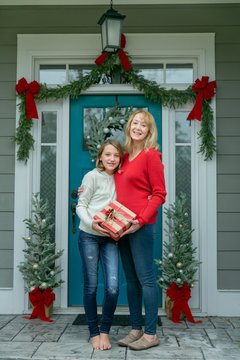 Mother And Daughter With A Christmas Gift Outside Their Home With A Festive Decorated Front Door