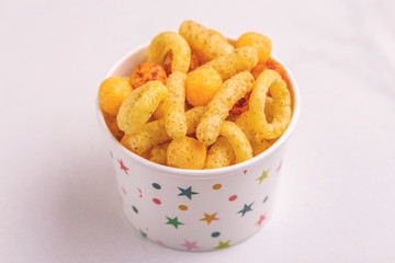 Assorted puff corn snacks in paper cup on light background