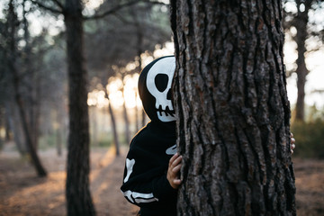 Kid in skeleton costume behind the tree in forest.