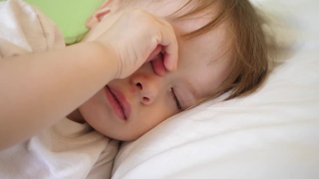 charming baby falls asleep on white bed in his bed in room at home. concept of sleeping child. child wants to sleep and rubs his eyes with his hands. kid sleeps in hospital ward. close-up