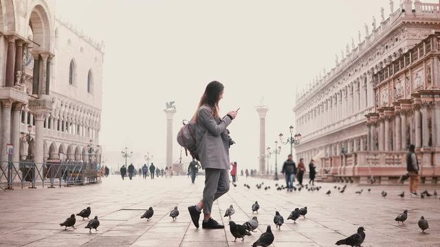 Wide shot of happy female tourist taking smartphone photos on amazing San Marco square full of birds in Venice, Italy.